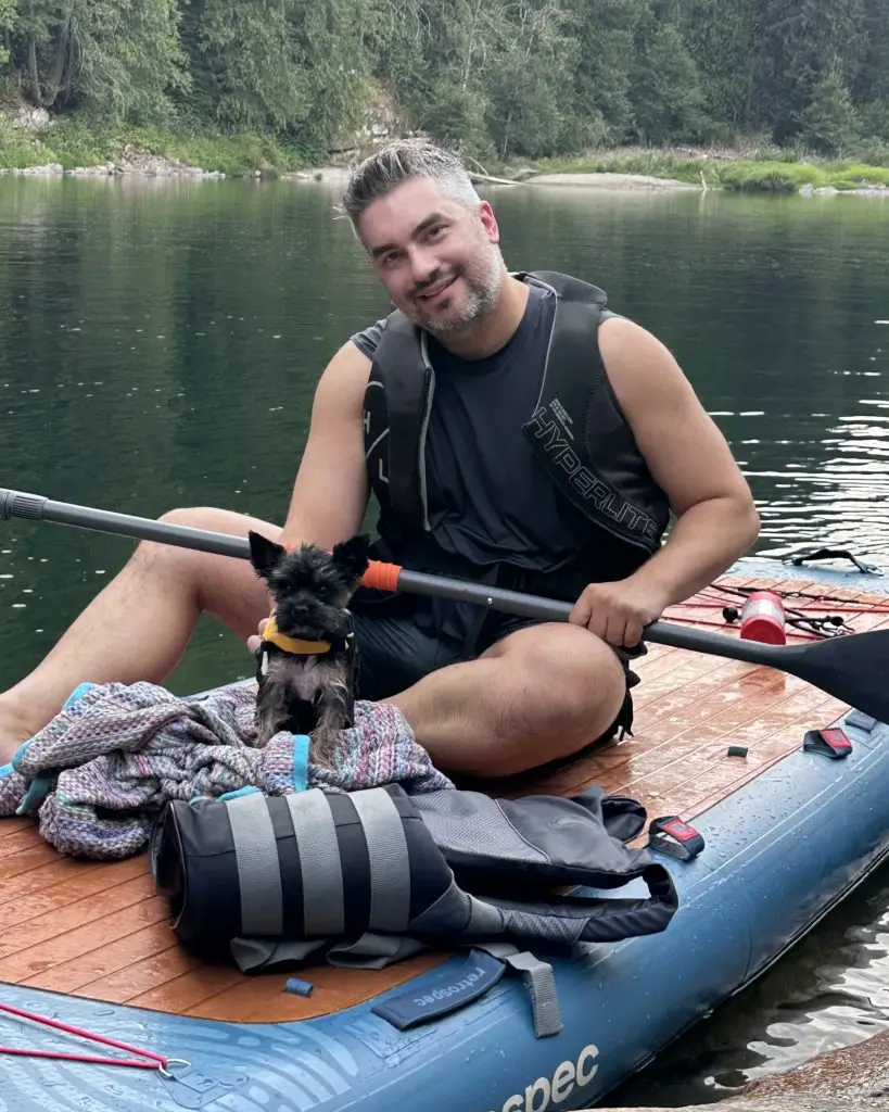 Smiling man in a life vest sits on a paddleboard with a small dog on a lake, forested shoreline in the background.