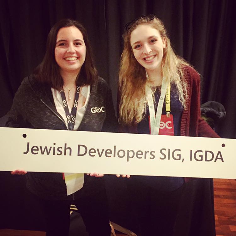 Two women smile as they hold a sign reading 'Jewish Developers SIG, IGDA' at a conference event.