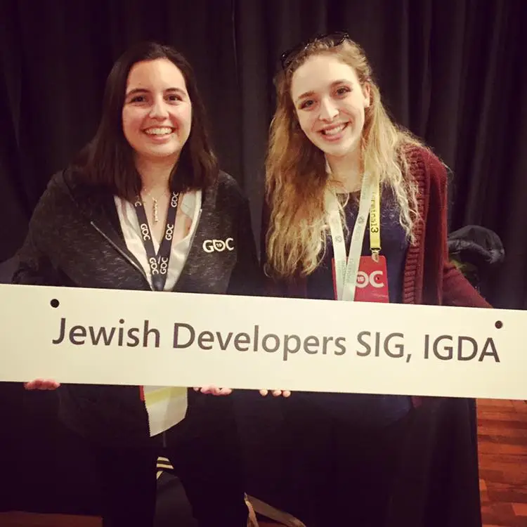Two women smile as they hold a sign reading 'Jewish Developers SIG, IGDA' at a conference event.