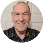 Close-up portrait of a smiling middle-aged man with short gray hair, wearing a blue plaid shirt against a light door background.