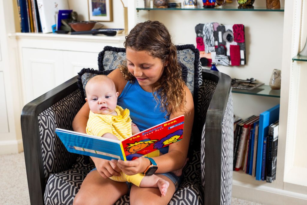 Woman reading a bright picture book to a baby while seated in a patterned chair at home.