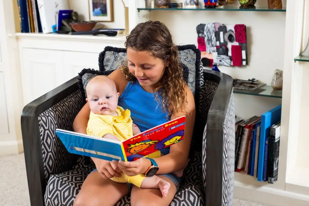 Woman reading a bright picture book to a baby while seated in a patterned chair at home.