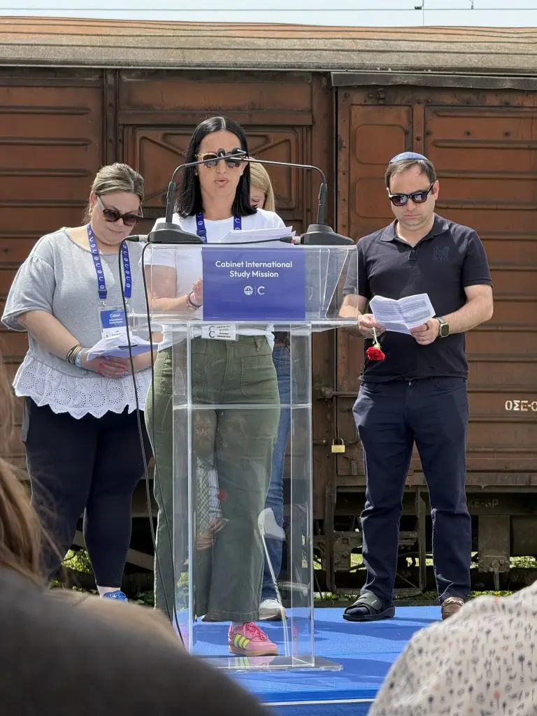 Center speaker at a clear lectern with a blue banner reading 'Cabinet International Study Mission'; two helpers hold papers on either side, rust-colored rail car in the background.