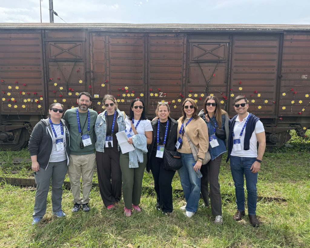 Group of eight adults posing in front of a rusted train car with decorative flowers and stars on sticks, outdoors on grass, wearing badges/lanyards.