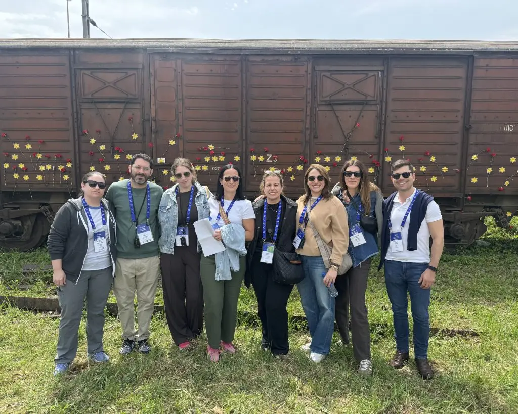 Group of eight adults posing in front of a rusted train car with decorative flowers and stars on sticks, outdoors on grass, wearing badges/lanyards.