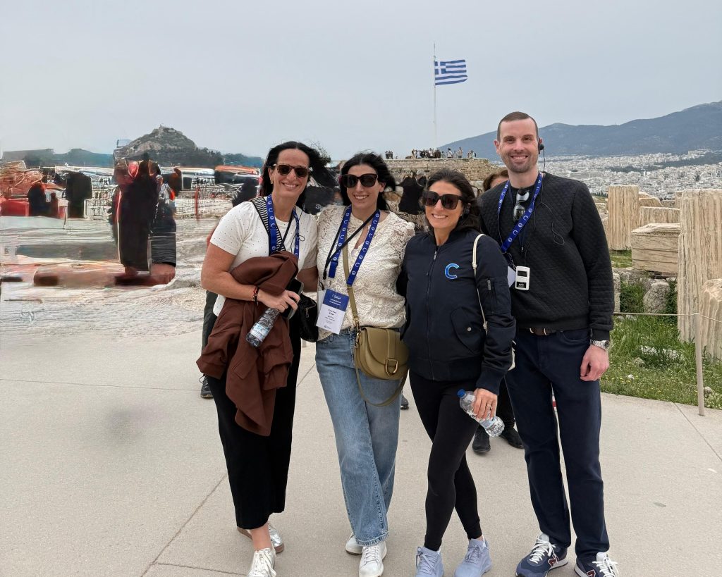 Four adults posing for a group photo at a scenic overlook in Greece, with a Greek flag and distant ruins in the background