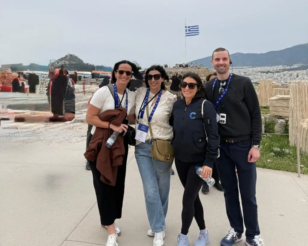 Four adults posing for a group photo at a scenic overlook in Greece, with a Greek flag and distant ruins in the background