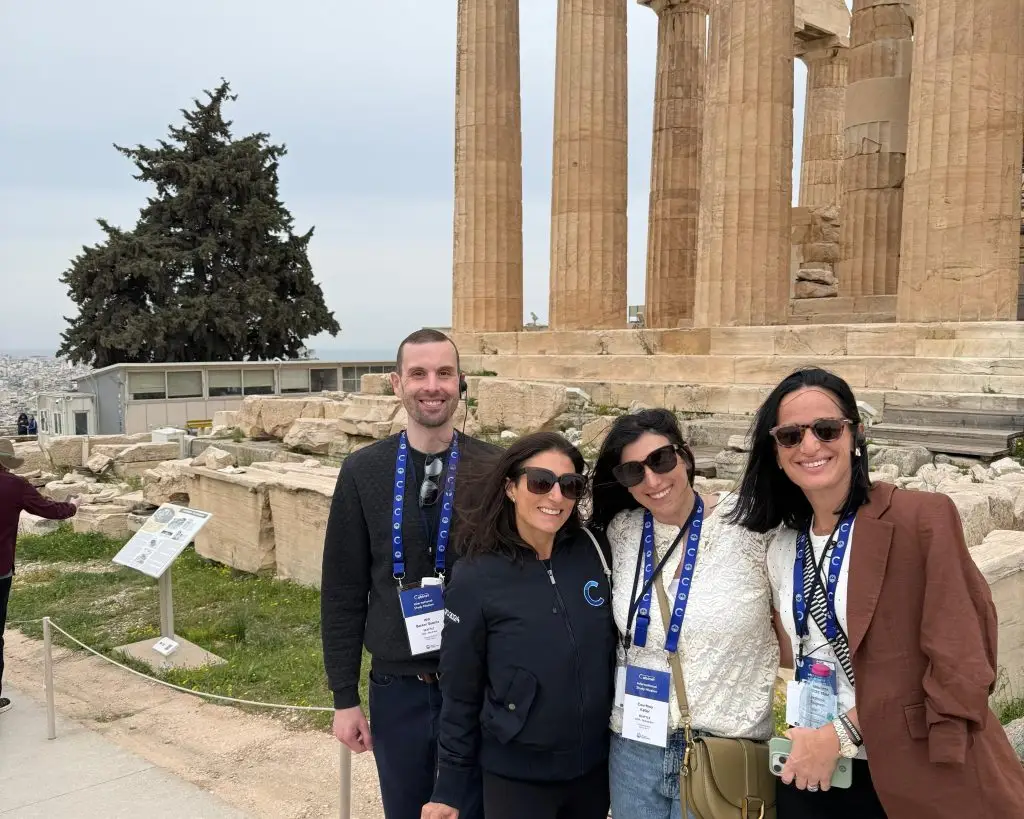 Group of tourists posing in front of ancient Doric columns at a historic ruin site, with a tree and sea in the background to the left.