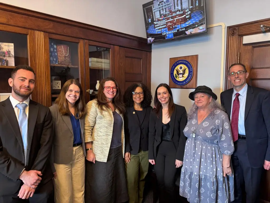 Group of professionally dressed people smiling in a doorway area near wooden cabinets and a U.S. House of Representatives seal on the wall.