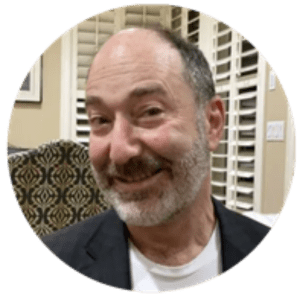 Close-up of a smiling man with a beard wearing a dark blazer indoors, with blinds in the background.