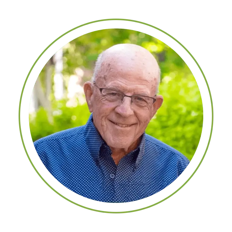 Smiling elderly man wearing glasses in a blue dotted shirt, outdoors with greenery.