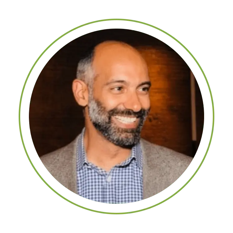 Smiling man with a beard in a beige blazer and checkered shirt, circular crop on green background.