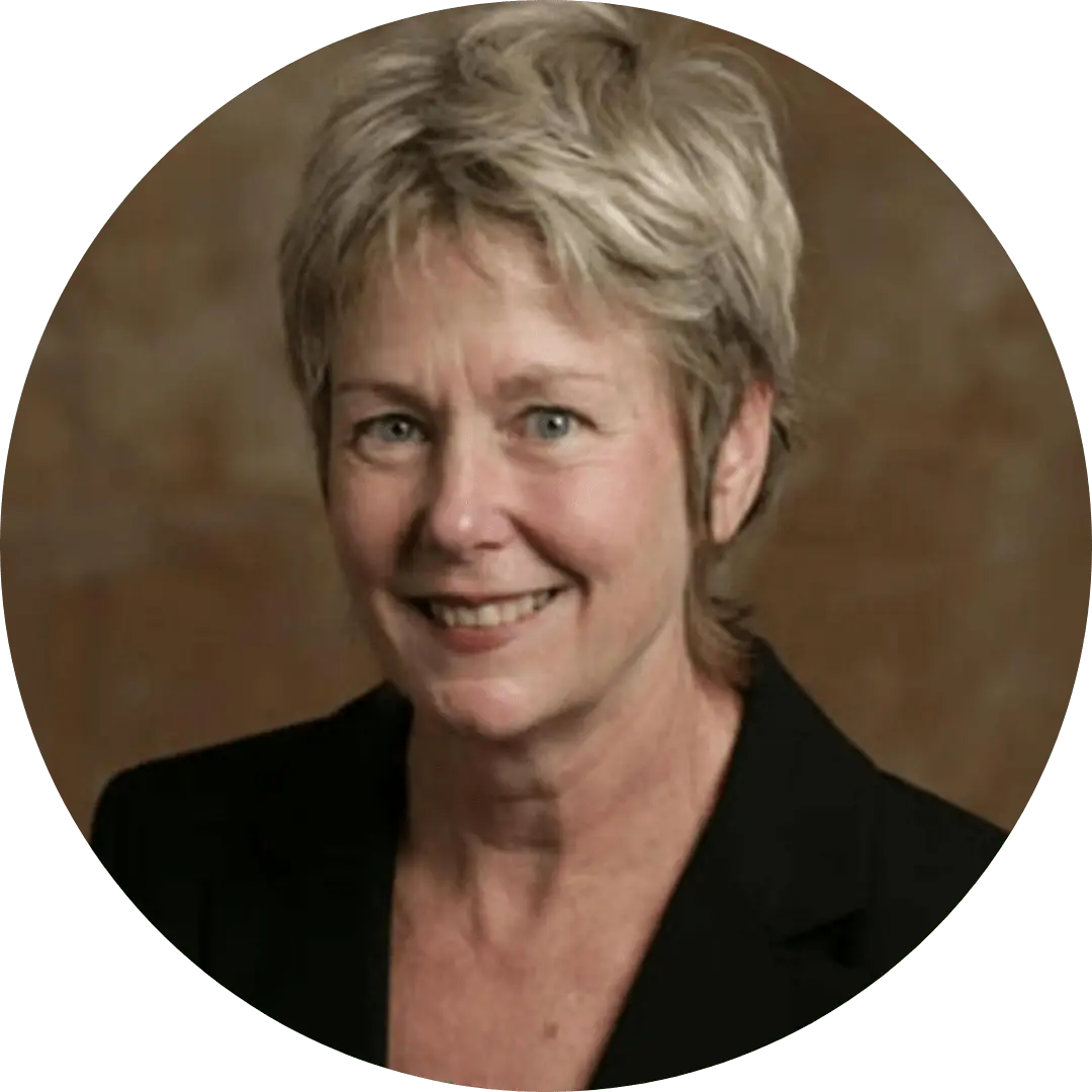 Smiling woman with short blond hair wearing a black blazer, facing the camera against a warm brown background.