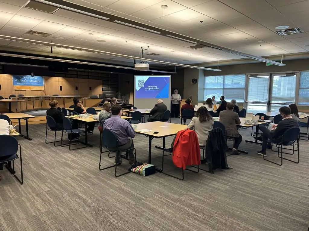 Audience seated at tables in a large conference room, presenter at a podium beside a projector screen showing 'Disrupting Anti-Jewish Ideas in the Current Climate' with a Seattle Public Schools banner in the back left.