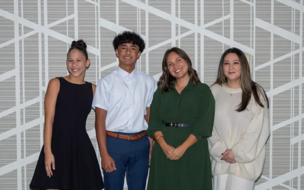 Four young adults standing side by side, smiling at the camera in front of a gray geometric patterned wall.