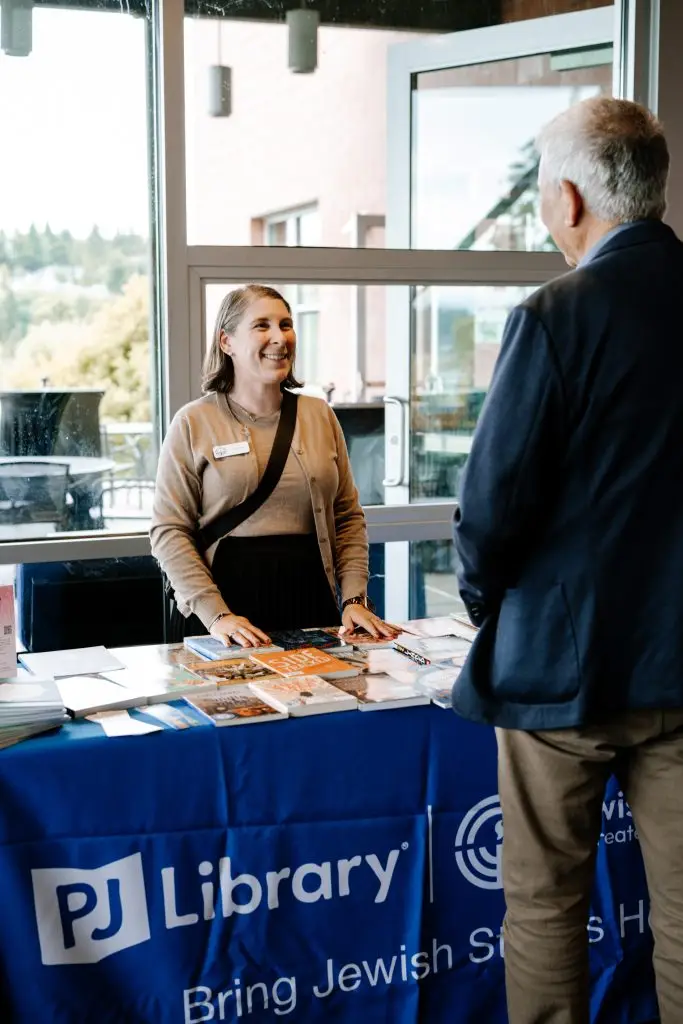 Smiling library staff member behind a desk with brochures, talking to a man by a glass door.