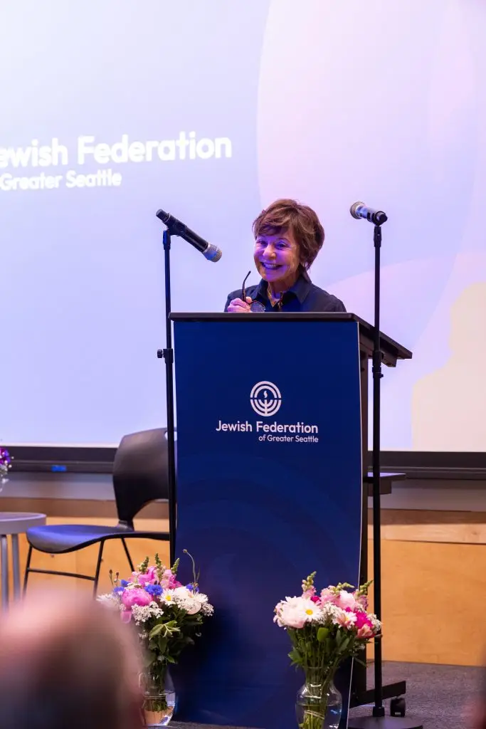 Smiling woman speaks at a lectern on stage, with two microphones and a Jewish Federation of Greater Seattle banner behind her, flowers in the foreground.