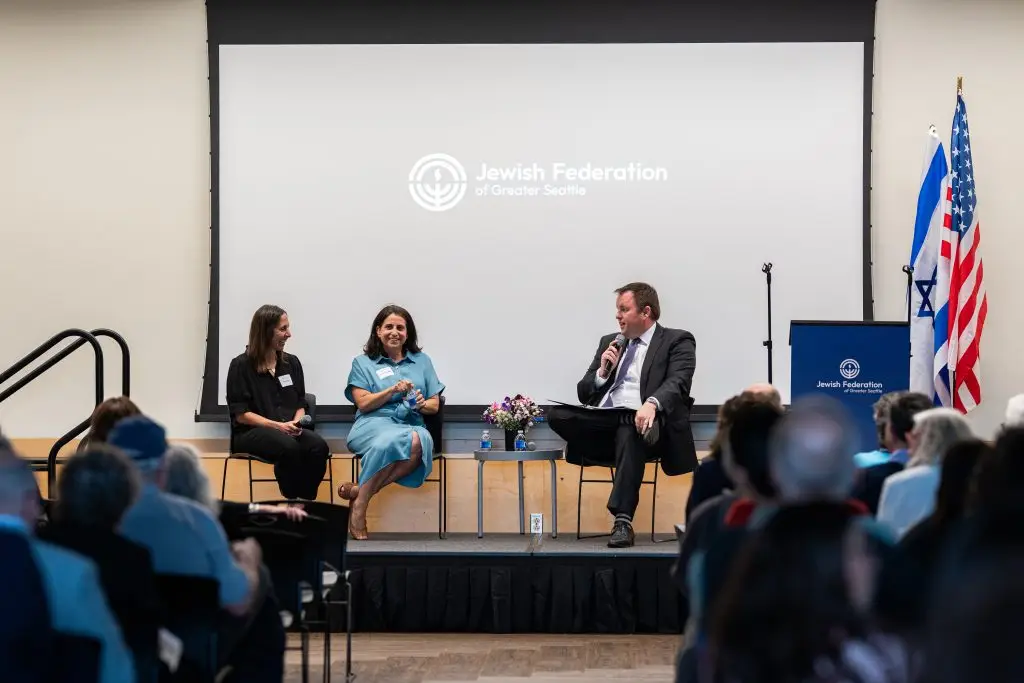 Panel discussion on a stage with three speakers; microphones, flowers, and a Jewish Federation of Greater Seattle backdrop visible, plus U.S. and Israeli flags to the right.