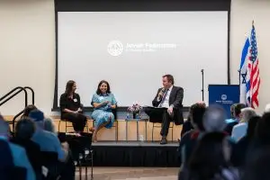 Panel discussion on a stage with three speakers; microphones, flowers, and a Jewish Federation of Greater Seattle backdrop visible, plus U.S. and Israeli flags to the right.