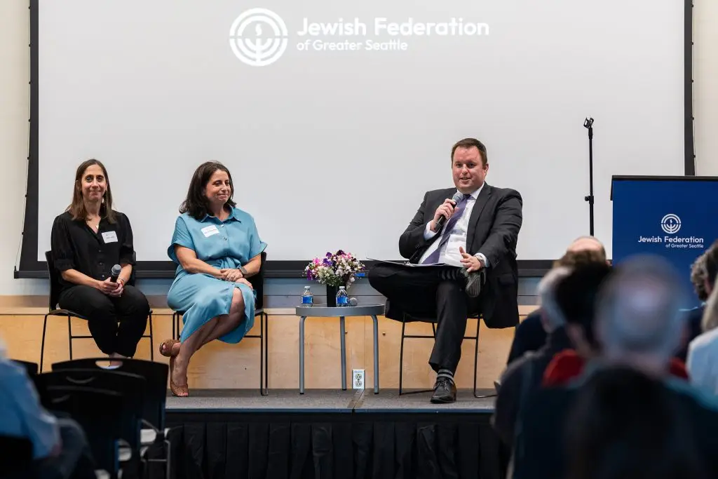 Panel discussion with three speakers on stage for Jewish Federation of Greater Seattle; man in suit holds microphone, two women beside him, audience in foreground.