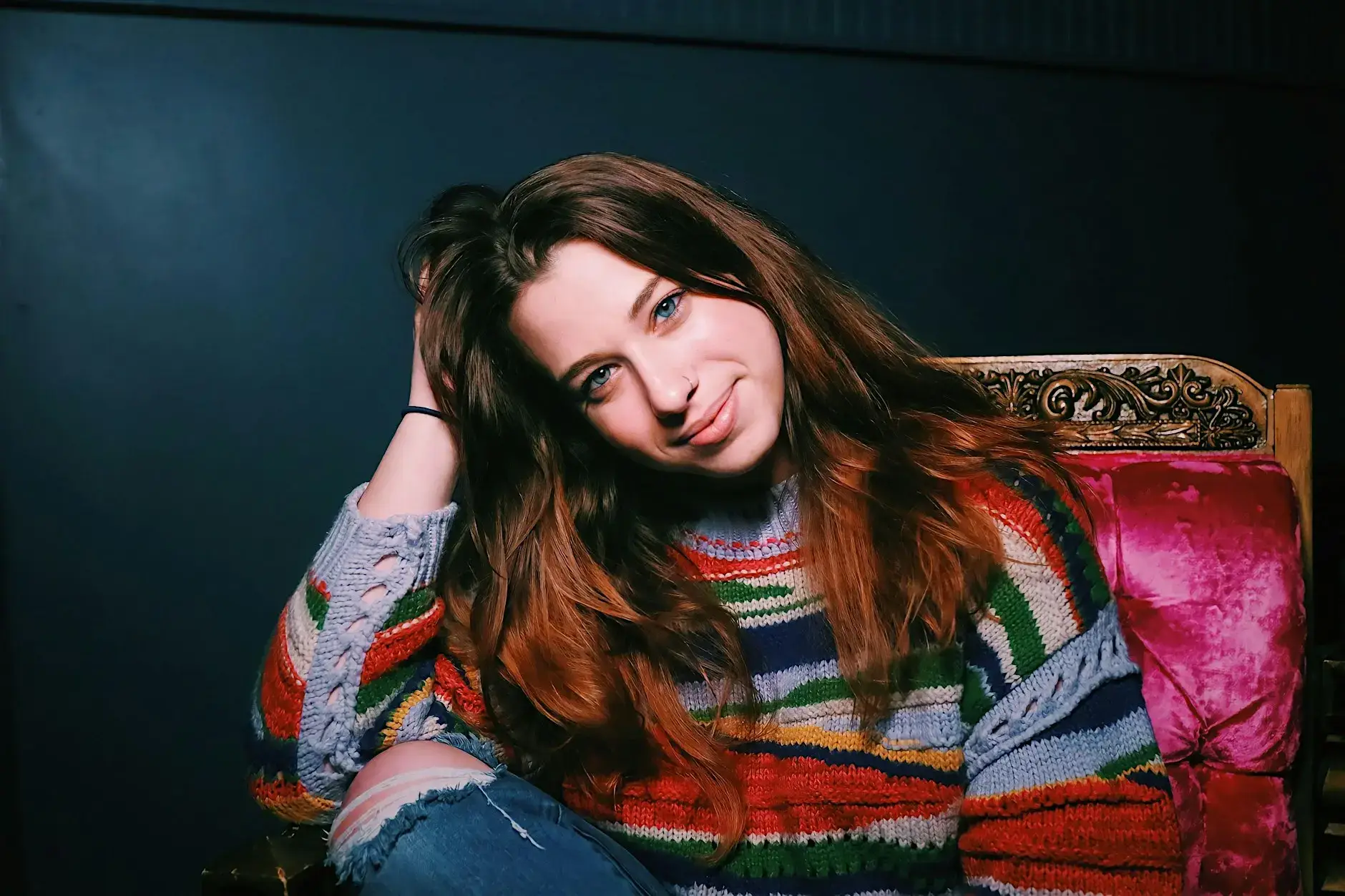 Young woman with long brown hair in a colorful striped sweater, smiling softly with her head tilted, seated on a carved wooden chair.