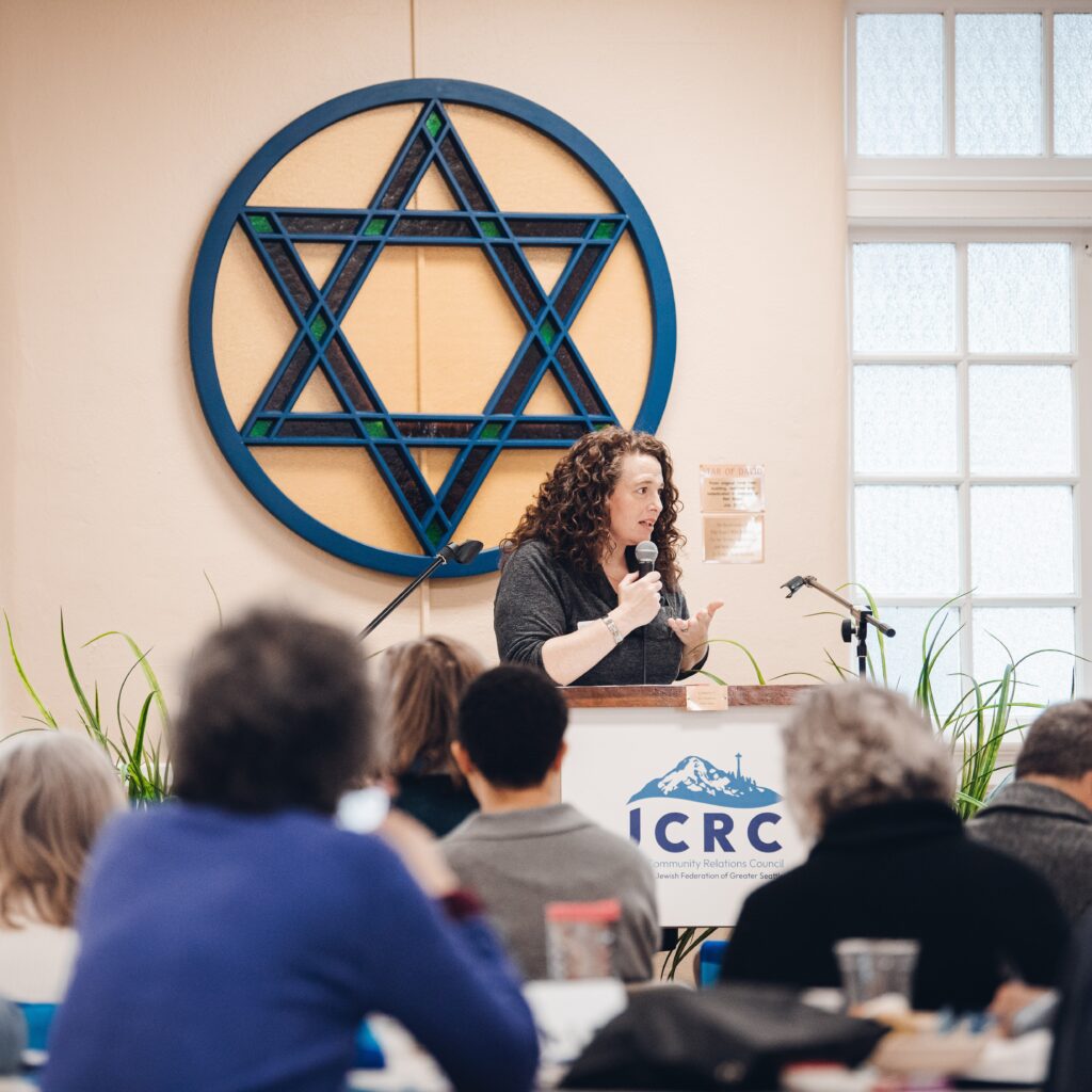 Woman speaking at a podium in front of a large blue geometric wall art and ICRC banner, addressing an audience.