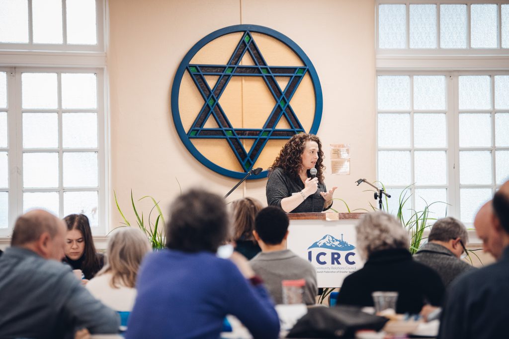 Female speaker with microphone at a podium in a room, audience watching, blue circular geometric wall emblem behind her.