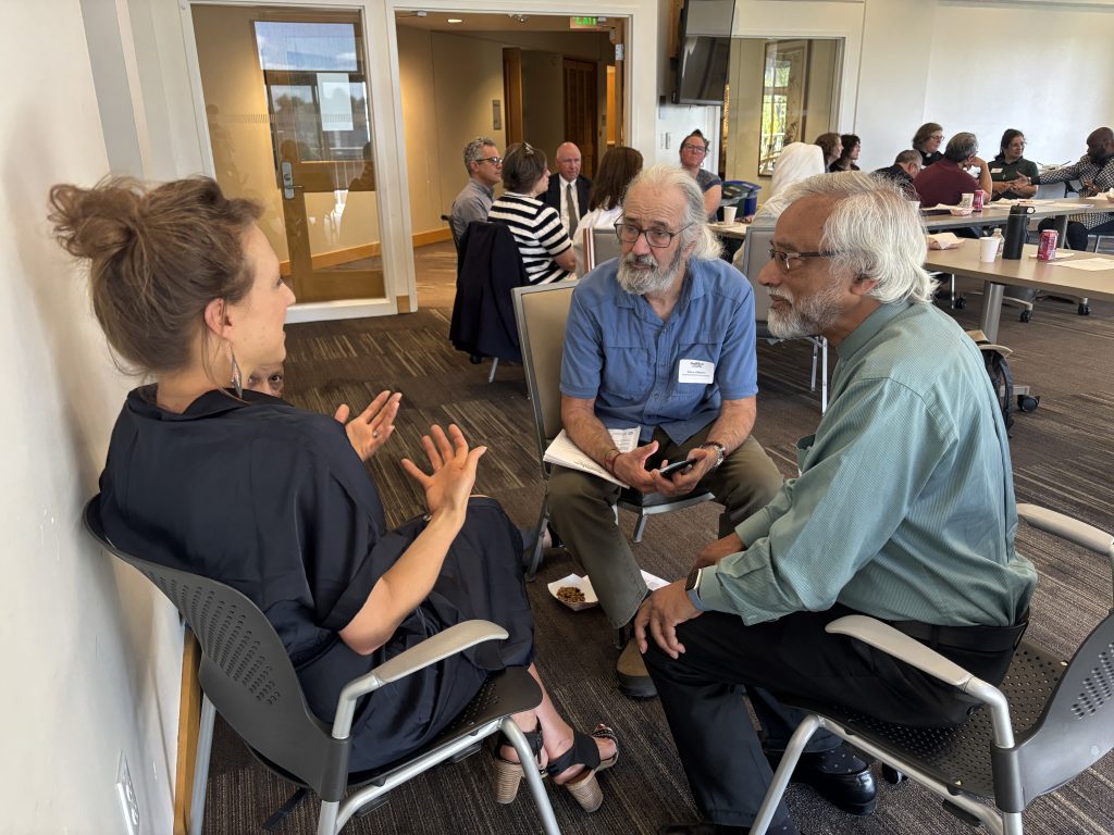Group of adults in a conference room engaged in conversation; two older men sit in the foreground while a woman on the left speaks with hand gestures.