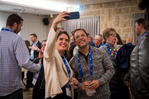 Smiling couple in front take a selfie at a conference, holding a phone overhead while others chat in the background with lanyards visible.