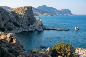 Rugged rocky coastline facing a calm blue sea, with small boats near the shore and distant hills on the horizon.