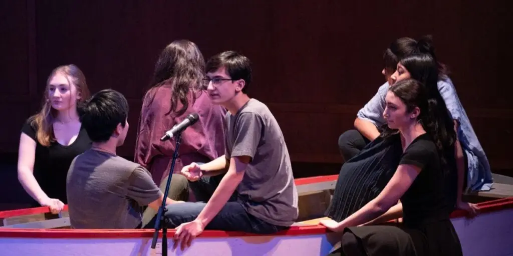 Six young adults sit in a red-trimmed wooden boat on a stage, chatting as if in rehearsal, with a microphone nearby.