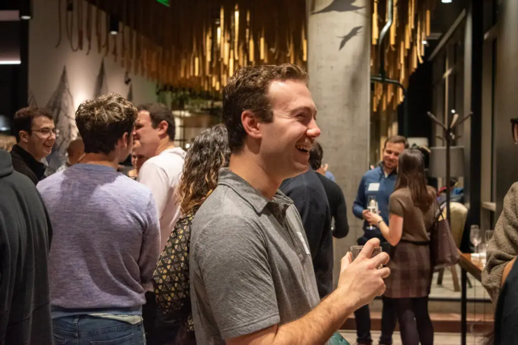 Smiling man in a gray shirt holding a drink at a crowded indoor social event, with wooden hanging decorations in the background.