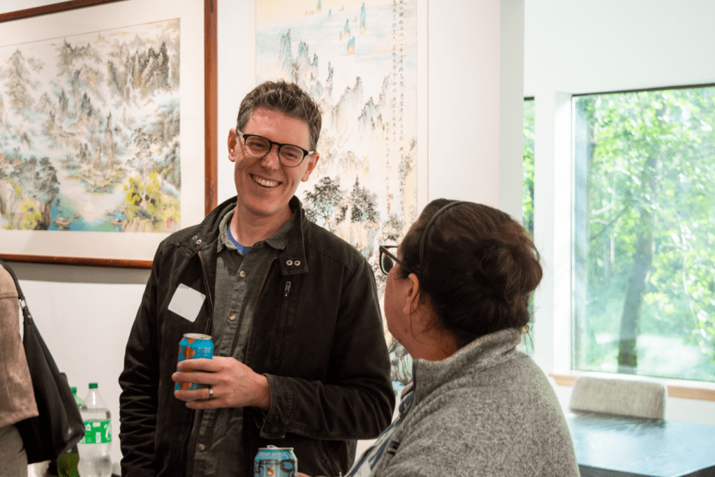 Smiling man with glasses wearing a dark jacket, holding a blue can, in an indoor art gallery with landscape paintings behind him.