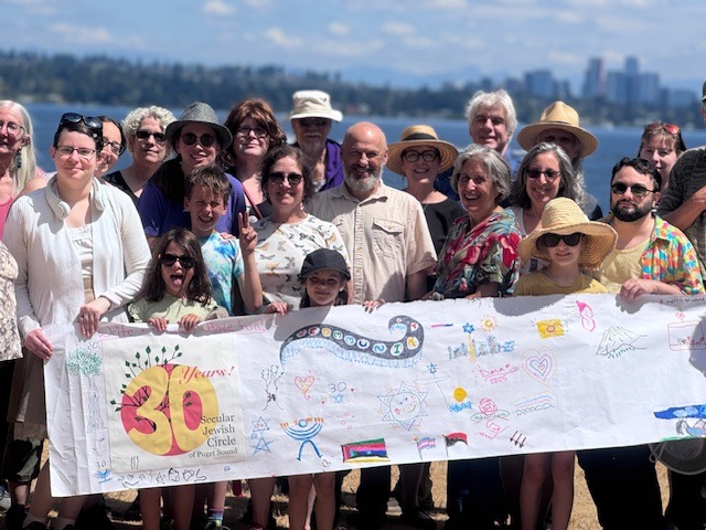 Group photo of about 25 people outdoors, smiling, holding a banner that says '30 years' for a secular Jewish circle, with water and city skyline behind.