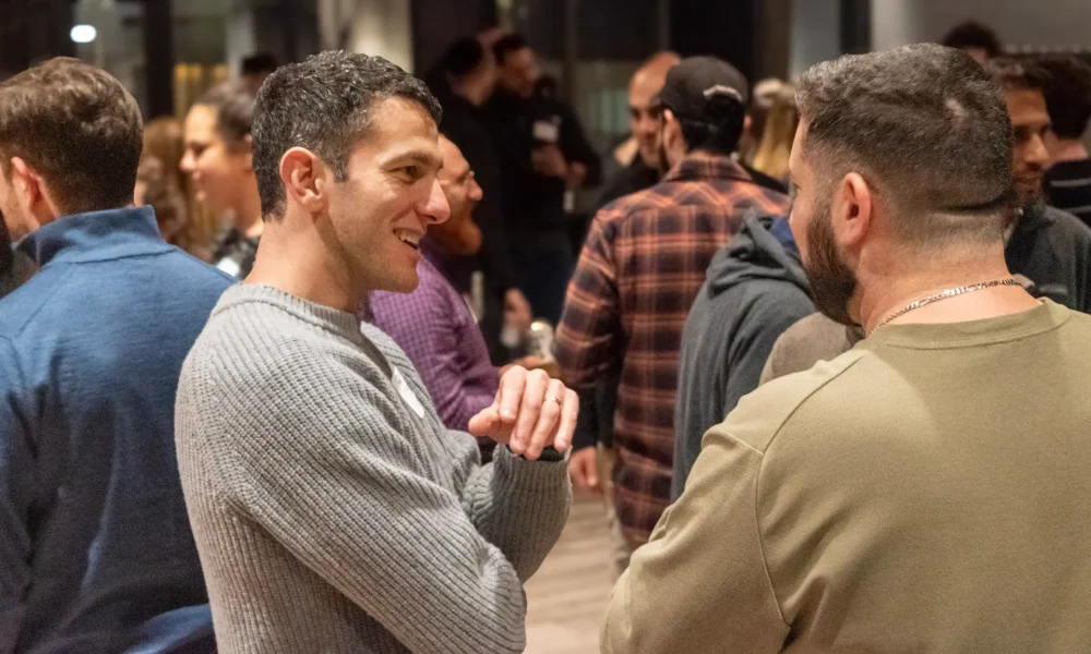 Two men in a friendly conversation at a crowded indoor event, one wearing a gray knit sweater and smiling at the other.