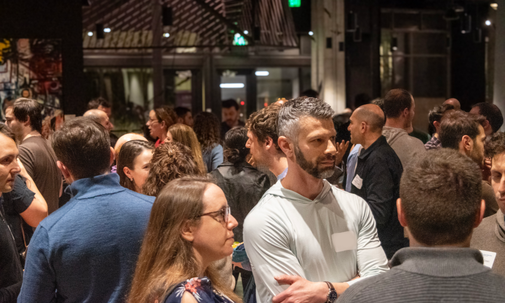 Crowded indoor social event with many people standing and talking in dim lighting.
