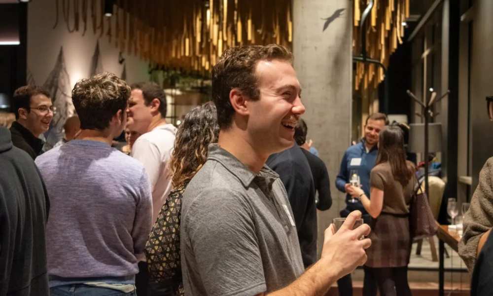 Smiling man in a gray shirt holding a drink at a crowded indoor social event, with wooden hanging decorations in the background.