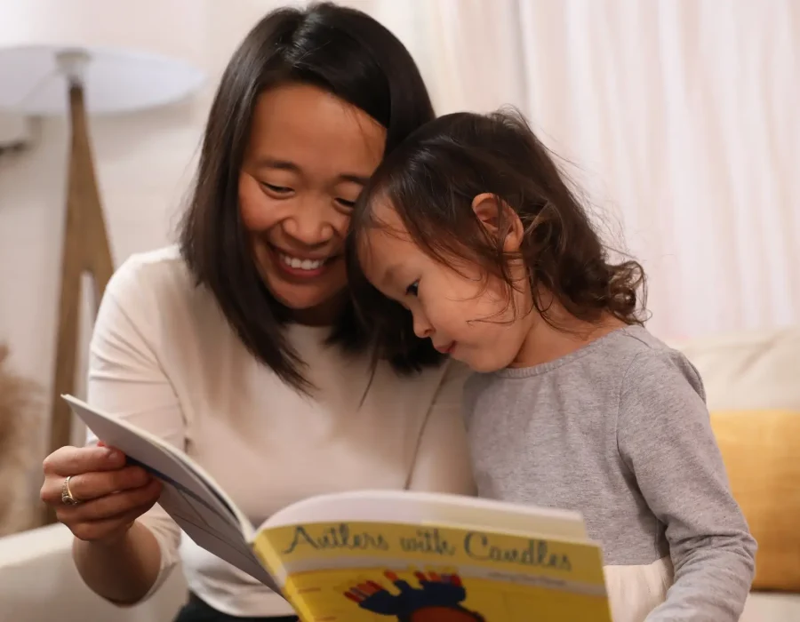 An adult woman and a child reading a colorful children's book together on a sofa, smiling and cuddling close.