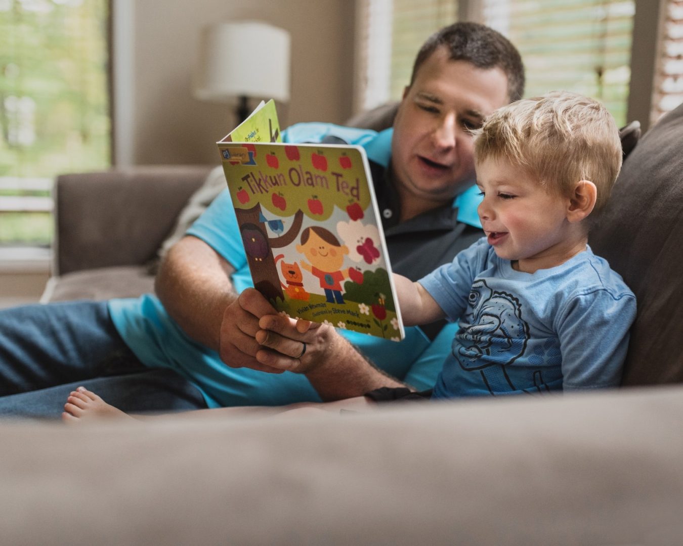 Father and young boy reading a colorful children's book together on a sofa.