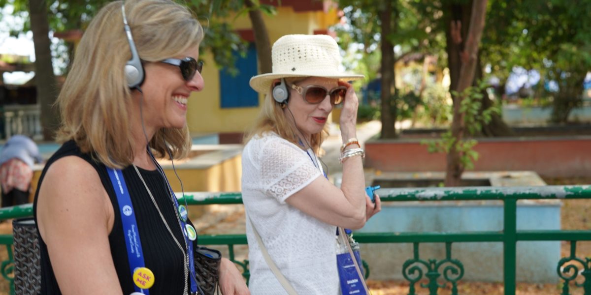 Two smiling women wearing headphones and lanyards chat outdoors beside a green decorative fence amid trees.