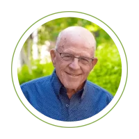 Smiling elderly man wearing glasses in a blue dotted shirt, outdoors with greenery.