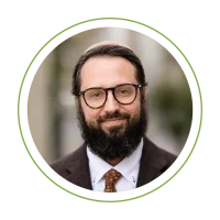 Portrait of a bearded man with glasses, wearing a brown blazer, white shirt, and patterned tie, smiling at the camera.