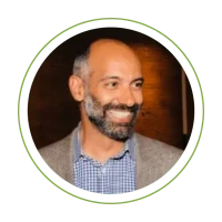 Smiling man with a beard in a beige blazer and checkered shirt, circular crop on green background.