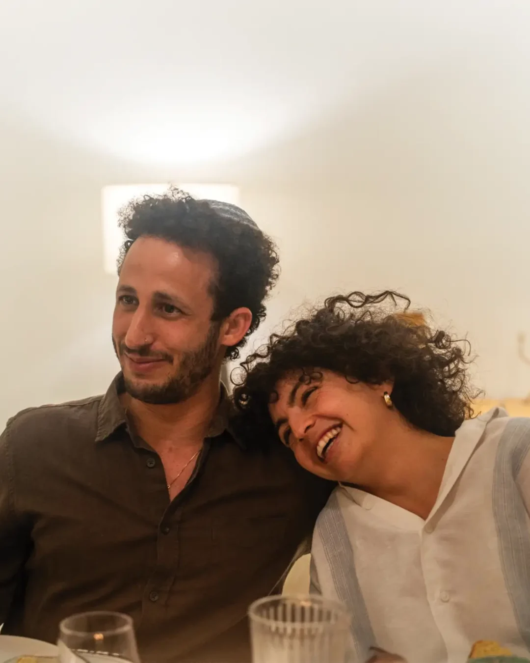 Two friends or a couple lean in close at a dining table, smiling warmly at the camera in a bright indoor setting.