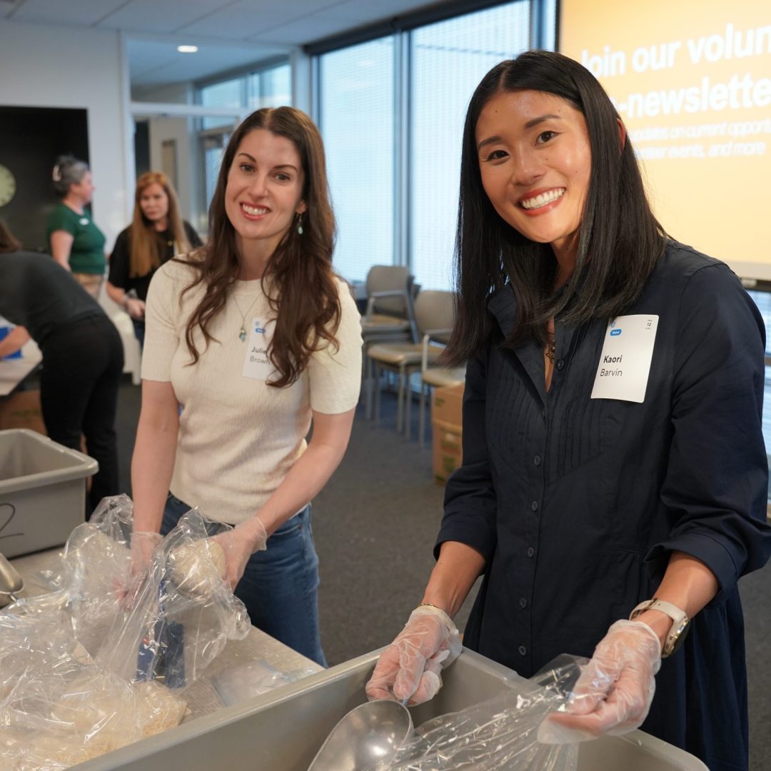 Two smiling volunteers wearing gloves pack food into a bin at a busy volunteer event on a sunny day.