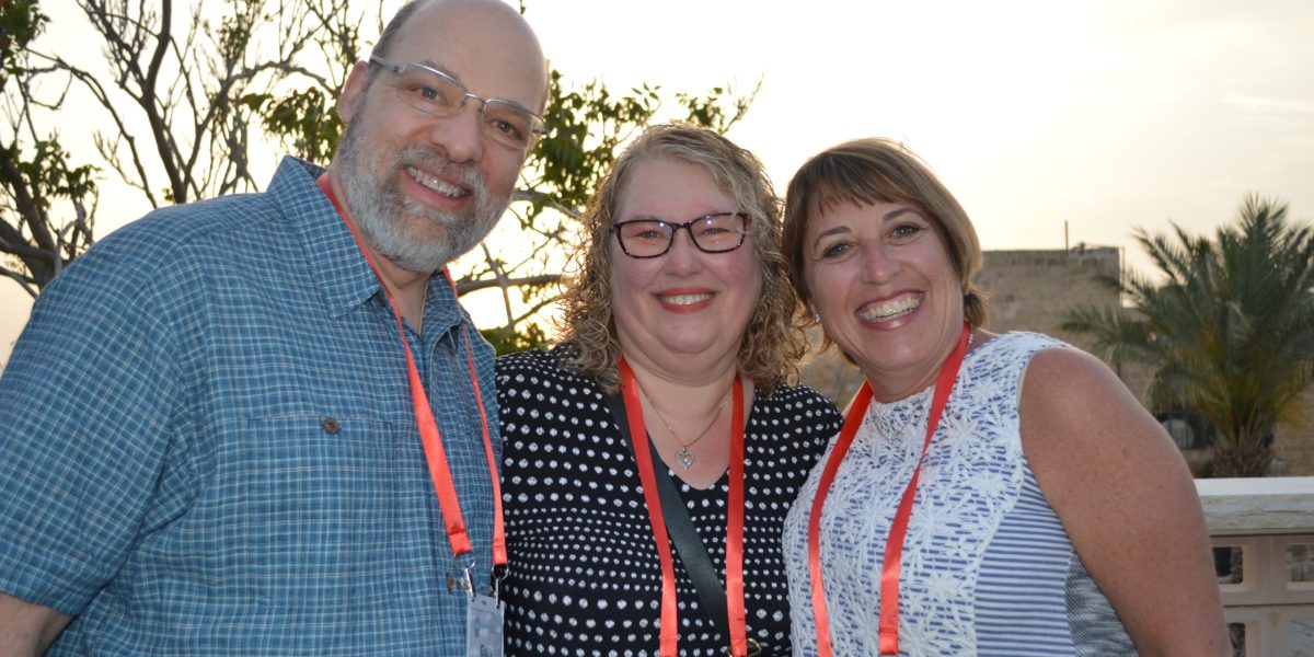 Three adults smiling for a selfie at an outdoor event, wearing name badges and red lanyards.