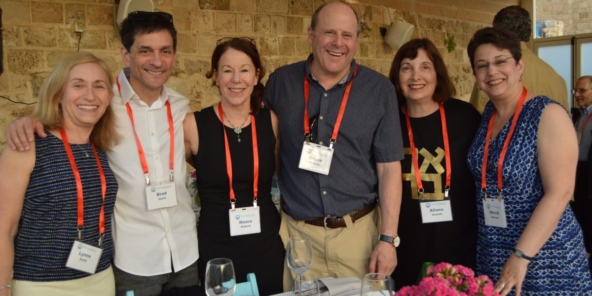 Six adults wearing name badges and red lanyards pose together at a table in a sunlit indoor event.