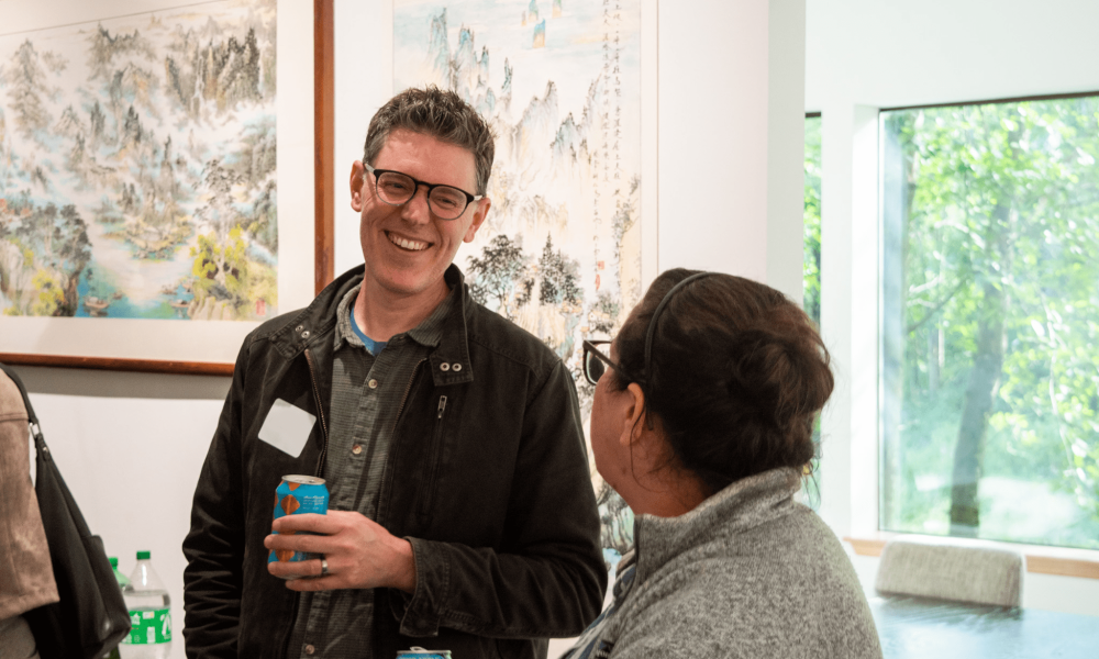 Smiling man with glasses wearing a dark jacket, holding a blue can, in an indoor art gallery with landscape paintings behind him.