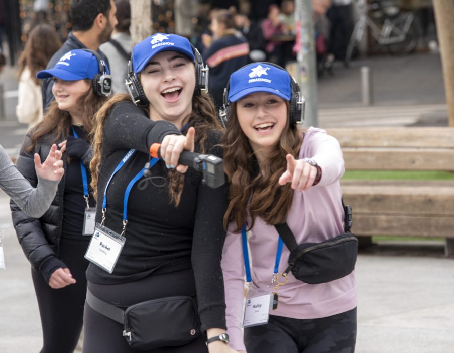 Three smiling young women in blue caps with headphones and event badges pose and point at the camera outdoors.