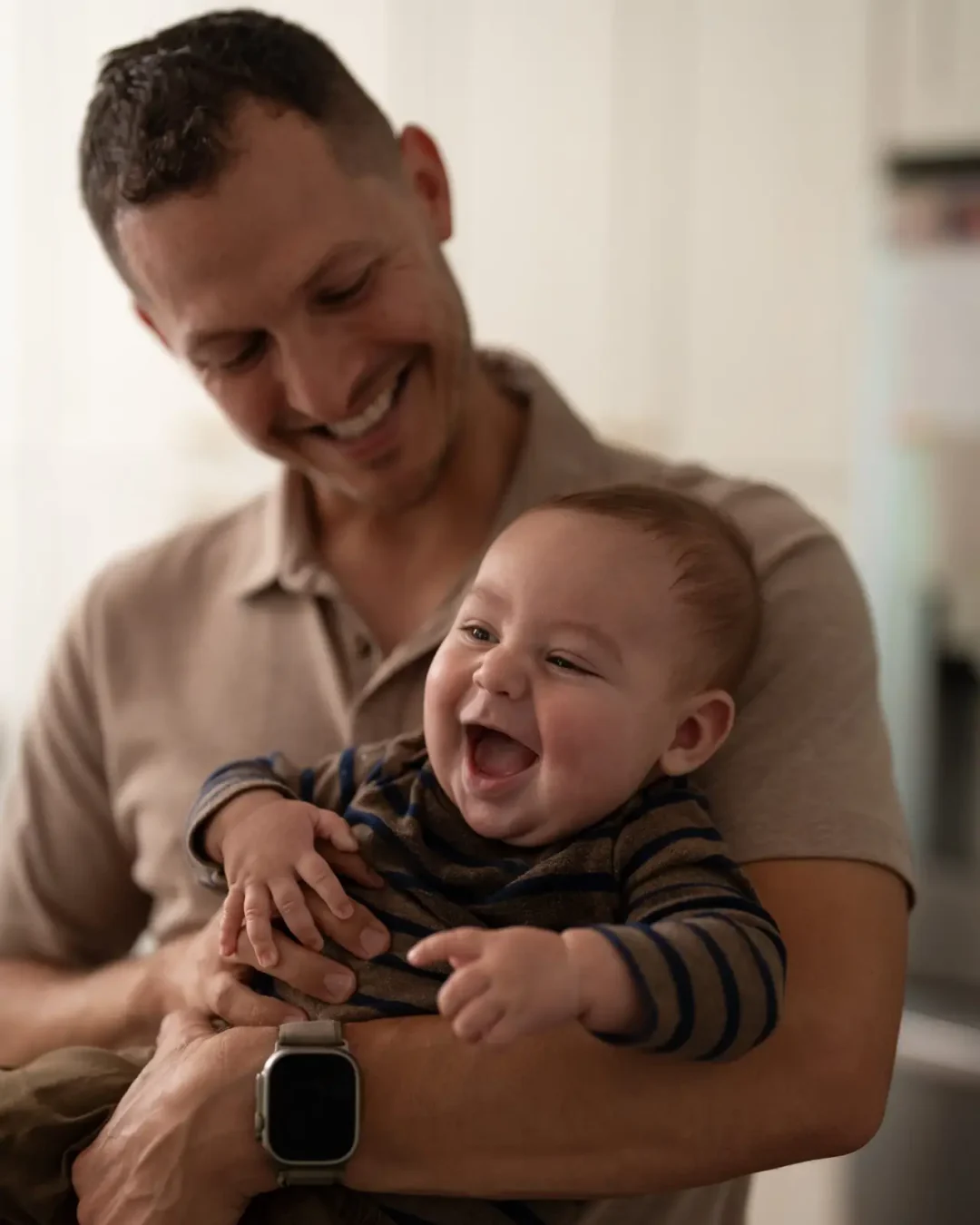Man smiling while holding a laughing baby in his arms indoors, both enjoying a moment together in soft light.
